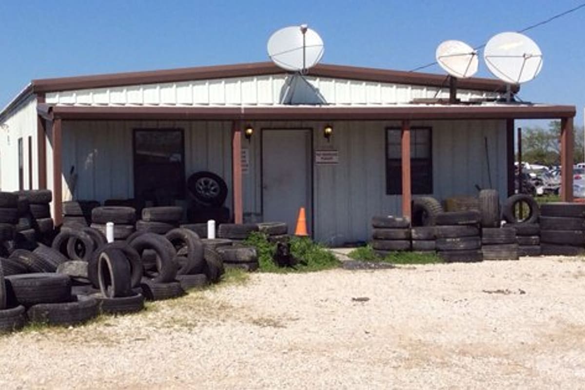 AAA Auto Salvage: Metal office building with satellite dishes and a sign, with large piles of used tires in the gravel yard in front