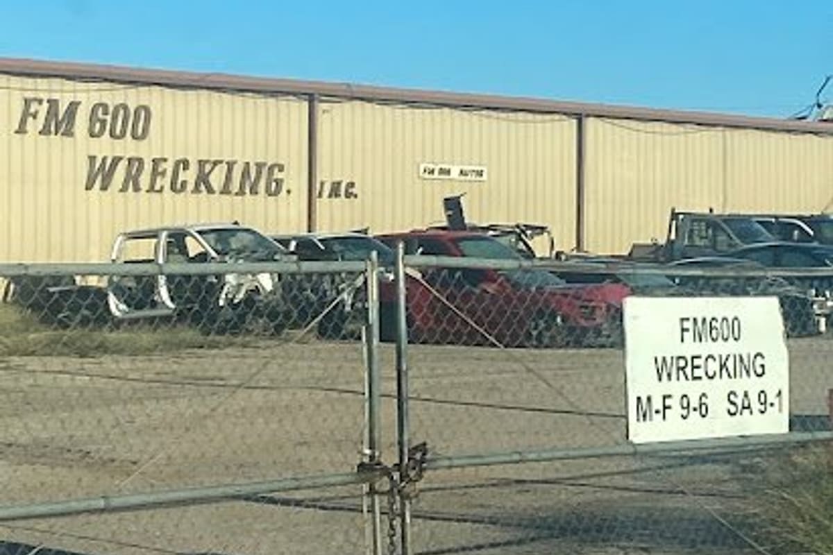 FM 600 Wrecking: Chain-link gate with hours sign in front of a tan metal building and rows of salvage vehicles under a clear blue sky
