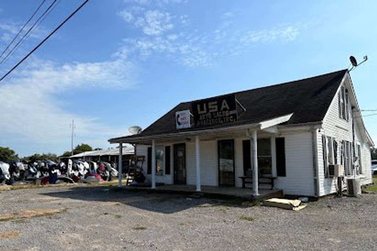 USA Auto Sales, Service & Salvage: White single-story office building with a covered porch and a rooftop sign, beside a gravel lot and stacked auto parts