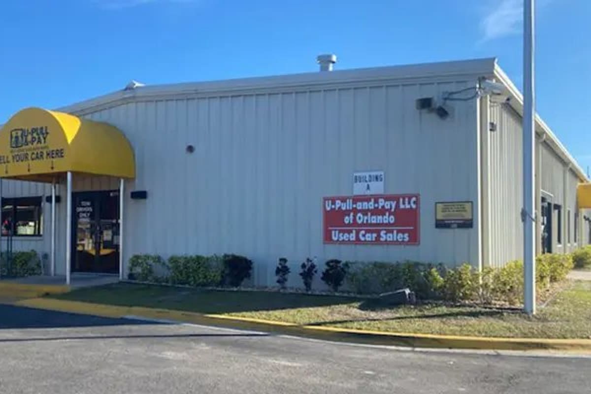 U-Pull-&-Pay Orlando: Beige metal building with yellow awnings over the entrance and a red sign, next to a paved driveway and landscaped strip