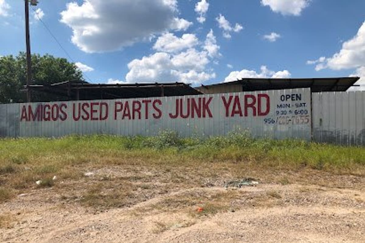 Amigos Used Parts Junk Yard: Painted white metal fence reading Used Parts Junk Yard with open hours, above a dirt lot and grass under a blue sky