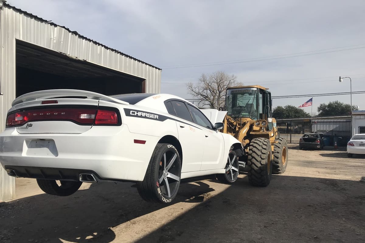 A1 Auto Partes: White Dodge Charger lifted by a yellow wheel loader in a dirt salvage yard beside a metal shed, with a flag in the background