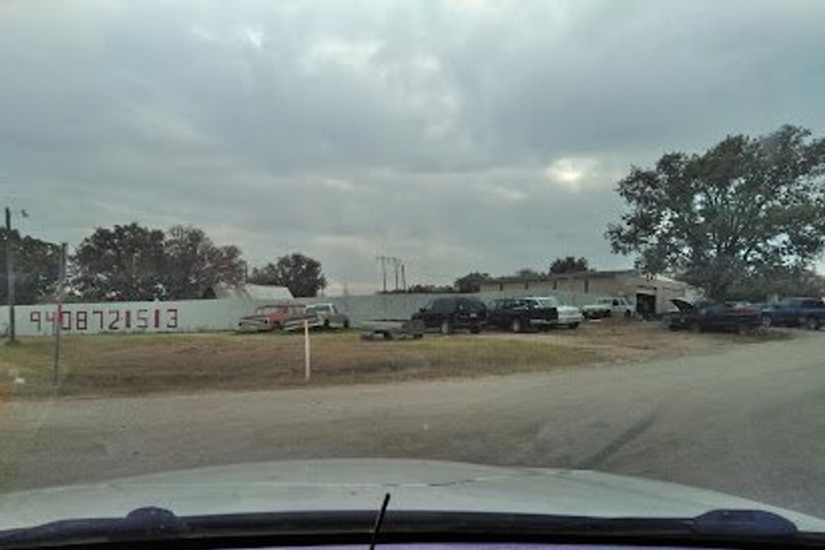 Phillips Auto Salvage: View across a gravel road toward a fenced salvage lot with trucks and cars parked in rows under a cloudy sky