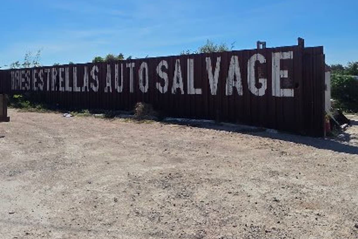 Tres Estrellas Auto Salvage: Long dark metal fence painted with large white lettering reading Tres Estrellas Auto Salvage beside a dirt lot
