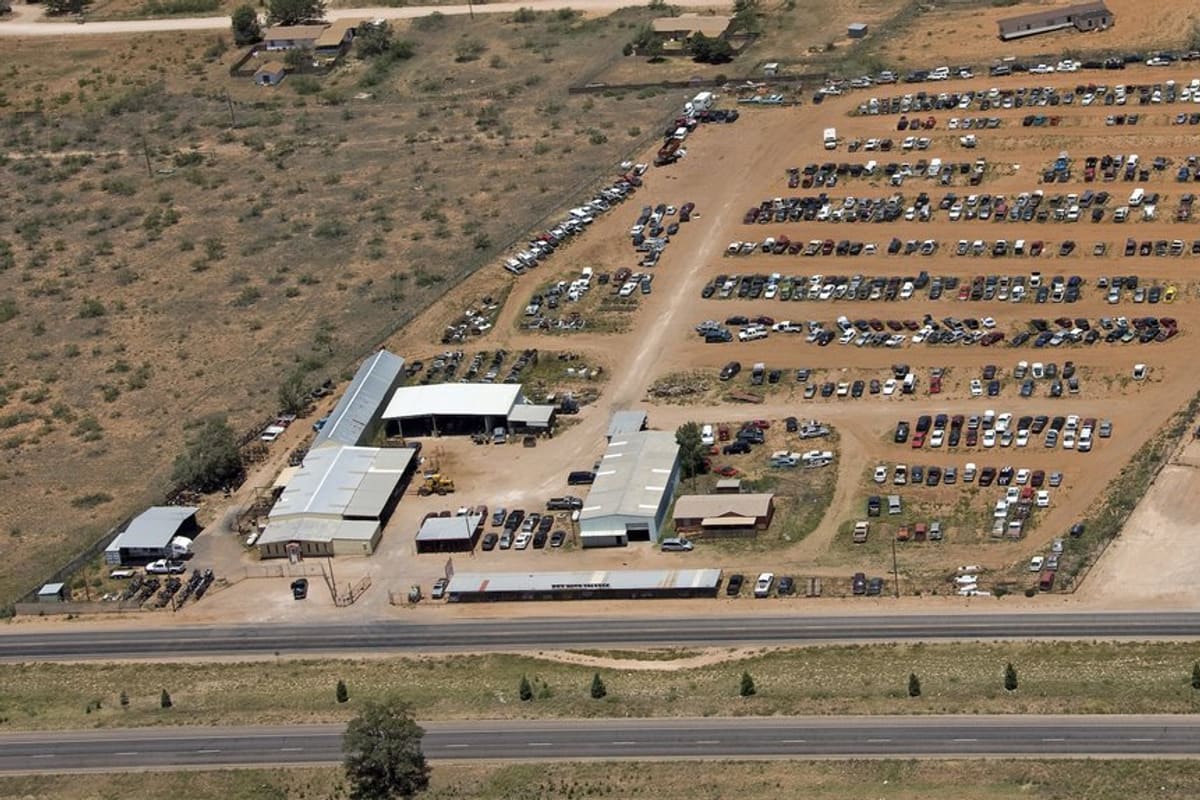 Box Auto Salvage: Aerial view of a large salvage yard with rows of cars on dirt lots, metal buildings, and a highway alongside