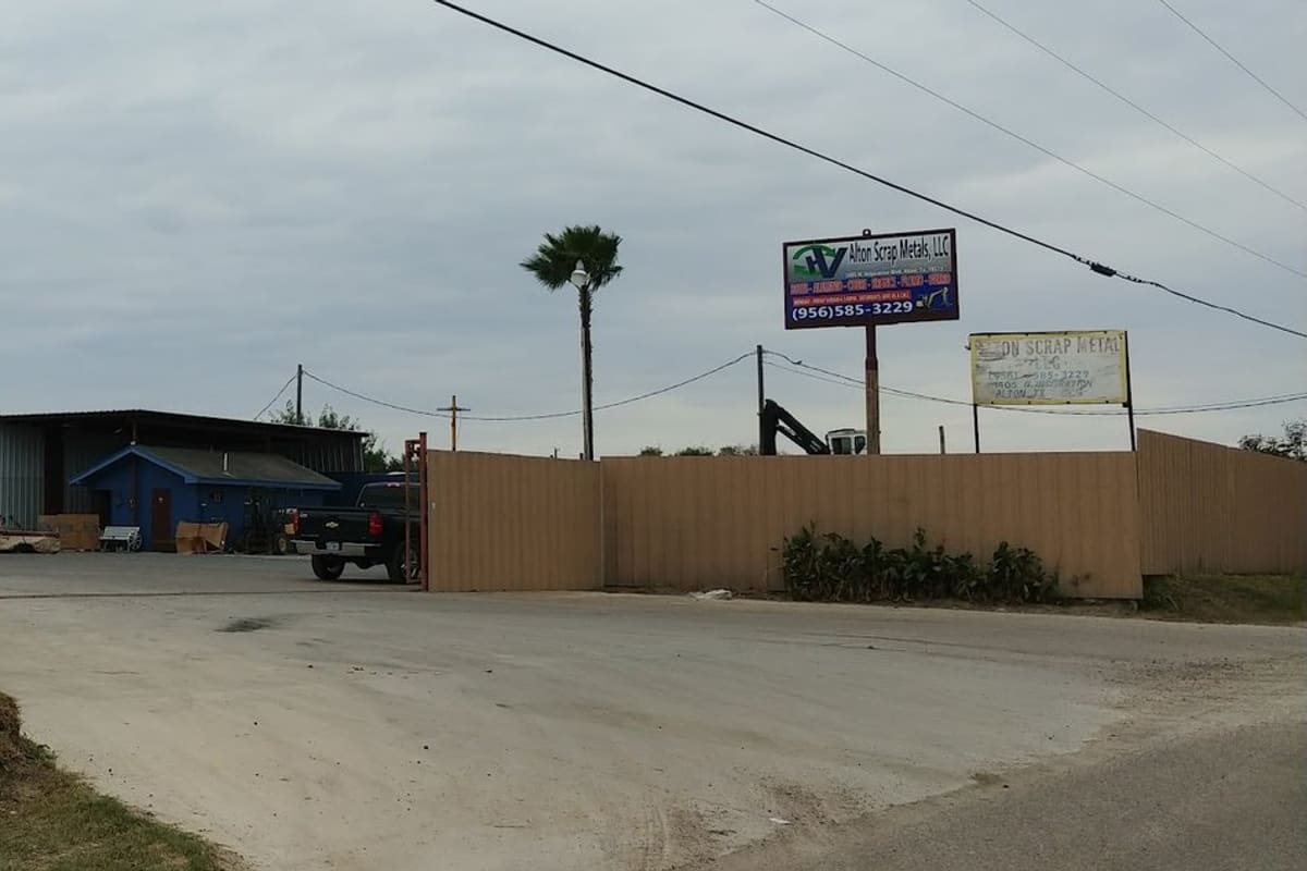 Alton Scrap Metal: Wide daytime view of a gated dirt driveway with tall tan metal fencing, a roadside sign and a blue shop building beyond