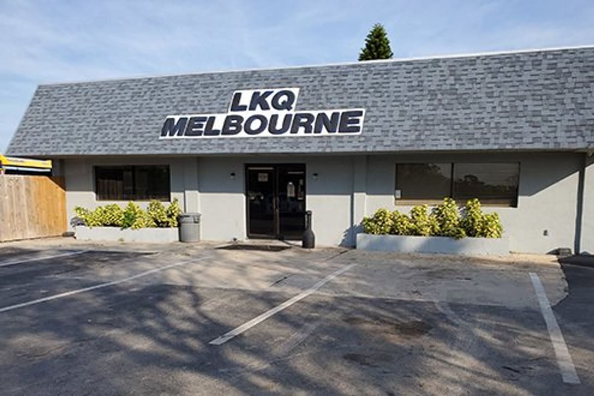 LKQ Melbourne: Single-story gray office building with shingled roof sign, planters, and empty parking lot in front