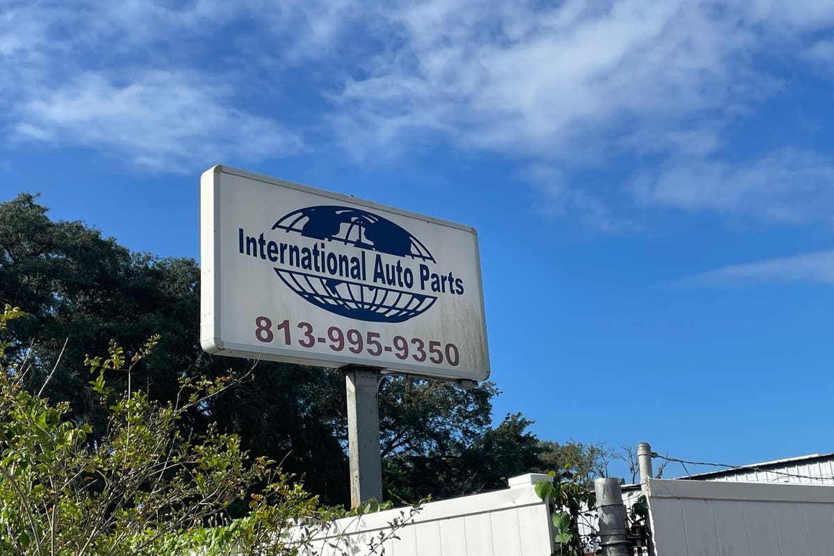 International Auto Parts Recycling: Roadside business sign with a globe logo and phone number on a pole against a blue sky, with a white fence below