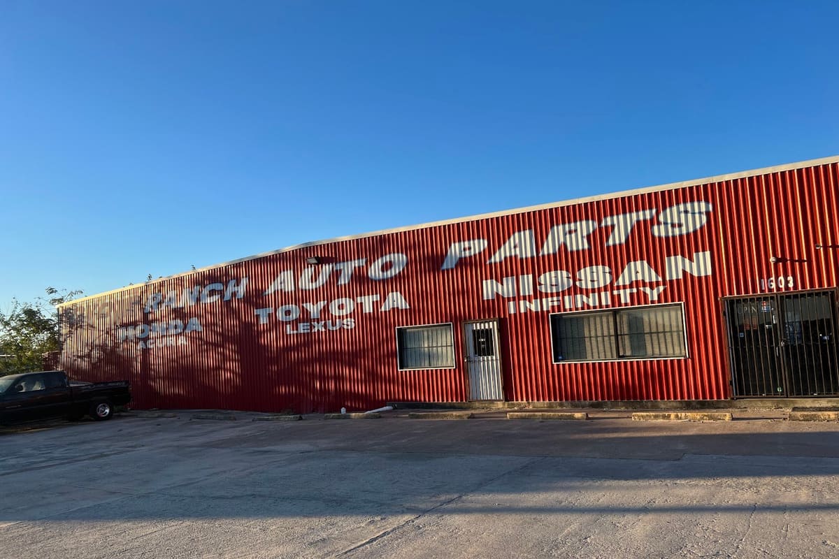 Ranch Auto Parts: Red corrugated metal auto parts building with large painted lettering listing car brands, beside an empty paved lot