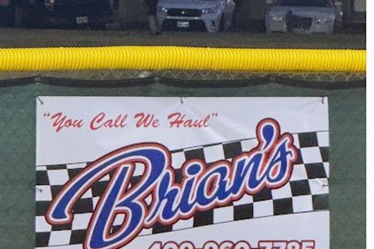 Brians Wrecker Service: Banner reading "You Call We Haul" with a checkered flag design and phone number, hung on a green fence at a ballfield