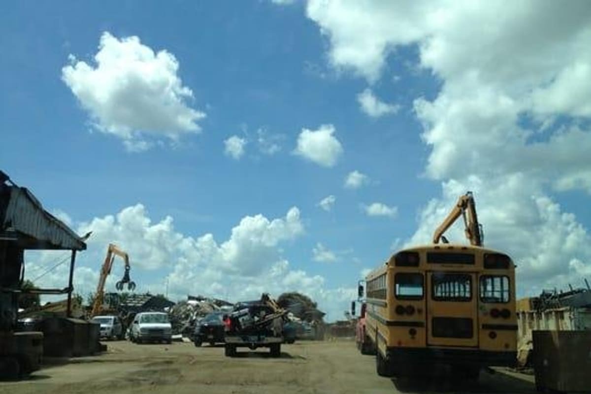 Basir Auto Recycling: Salvage yard with a yellow school bus, scrap piles and a crane with grapple under a blue sky