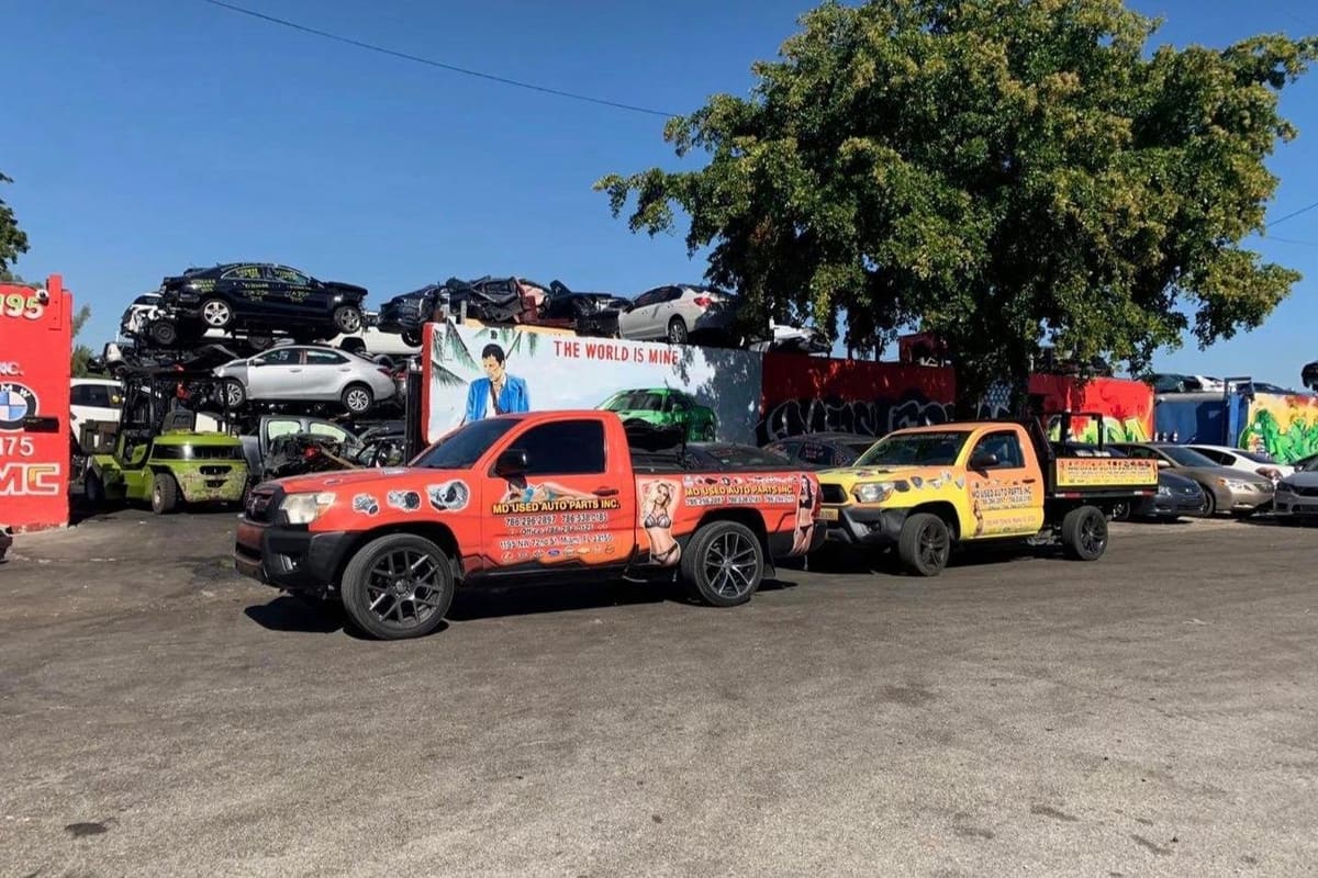 MD Used Auto Parts: Wide daytime view of a salvage yard with stacked wrecked cars, two decorated pickup trucks and a forklift on a paved lot