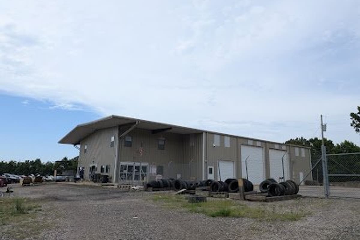 DIY Auto Parts: Large tan metal shop building with roll-up garage doors, stacked tires out front and a gravel lot under a cloudy sky