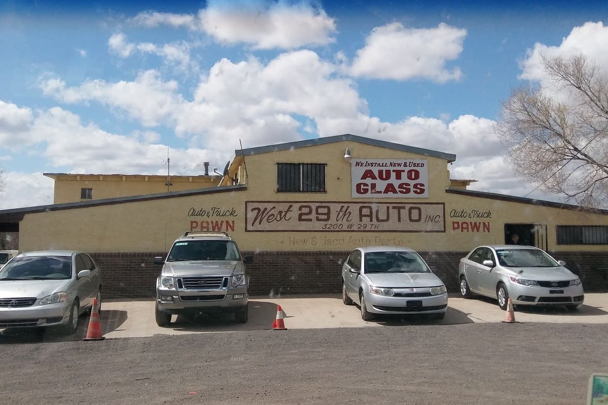 West 29 Auto: Yellow stucco shop building with painted wall lettering and an auto glass sign, with cars parked in front under a cloudy blue sky
