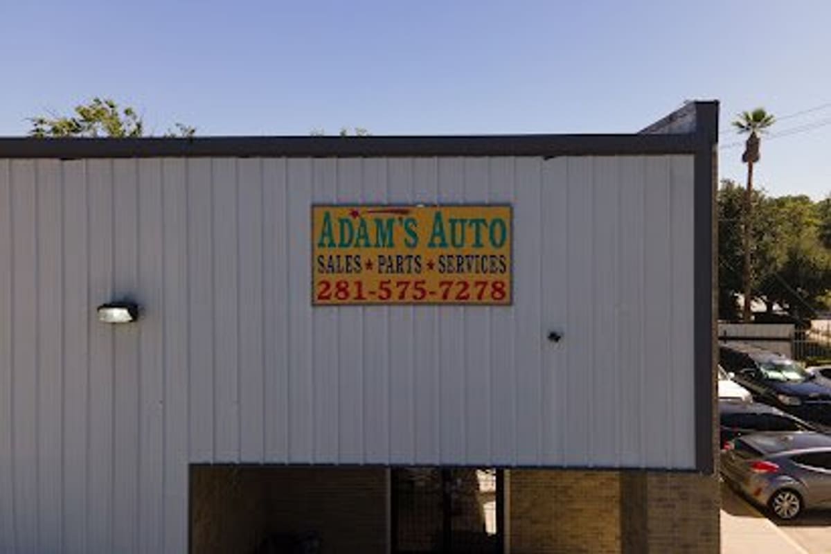 Adam's Auto Sales & Parts: White metal building with a painted sign reading sales, parts and services, with parked cars at the right edge