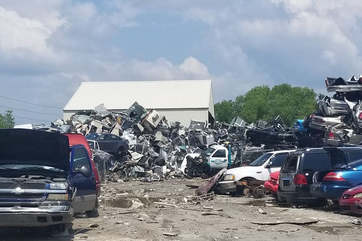 Express Recycling Group: Wide view of a muddy salvage yard with piles of scrap metal, stacked crushed cars and vehicles under a partly cloudy sky