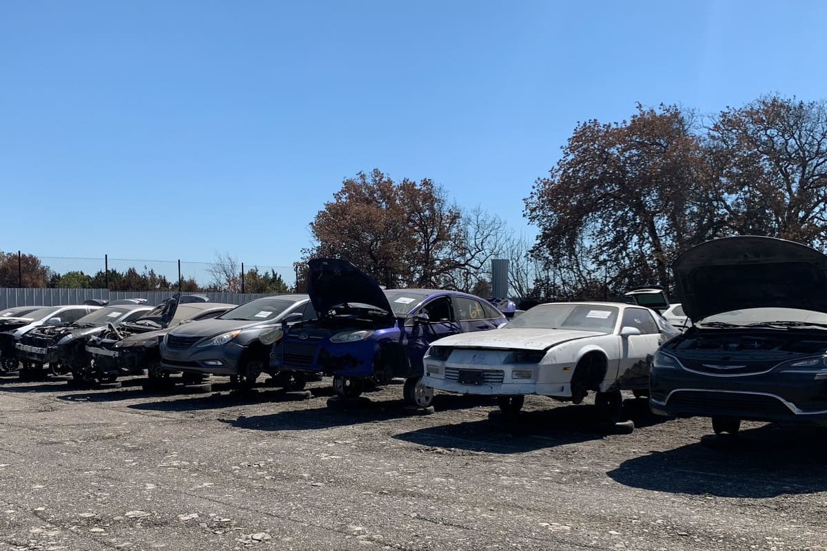 Budget Wrench A Part: Row of stripped cars with open hoods resting on stands in a gravel yard under a clear blue sky