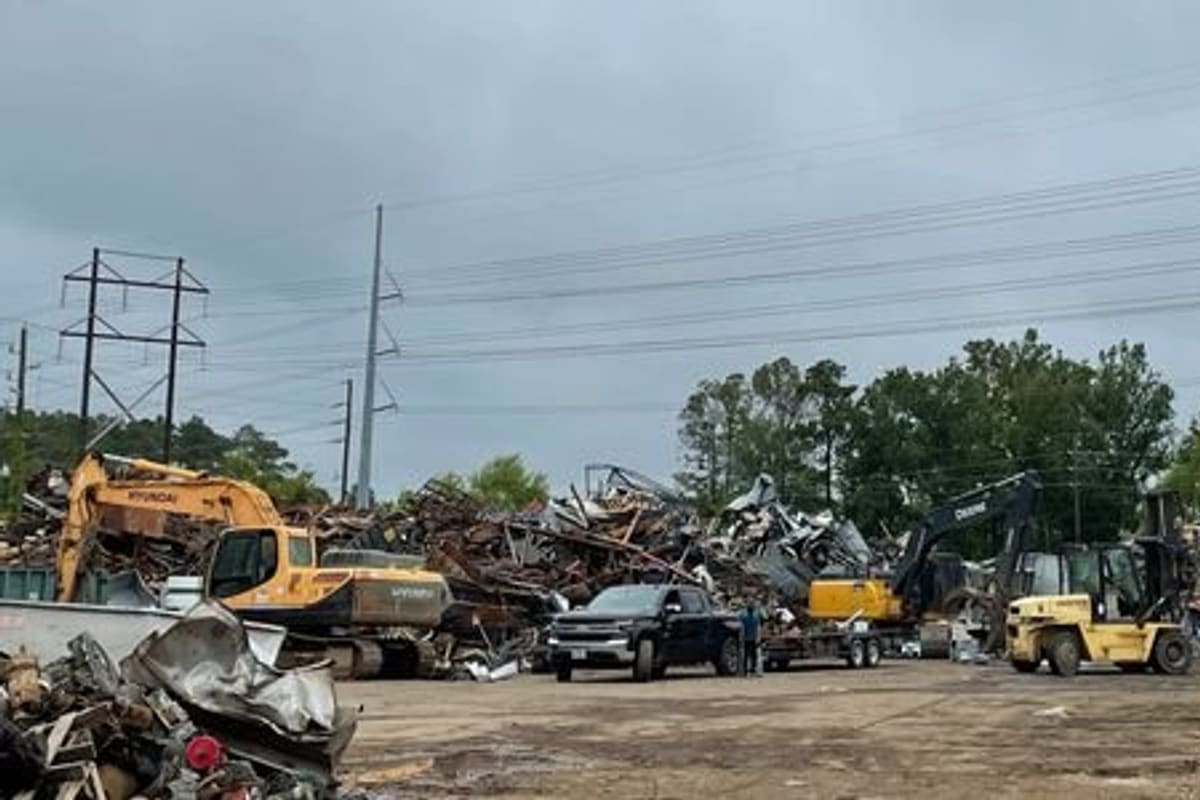 New Caney Recycling: Scrap metal yard with large piles of salvage, excavators, a forklift, and a pickup truck under cloudy skies