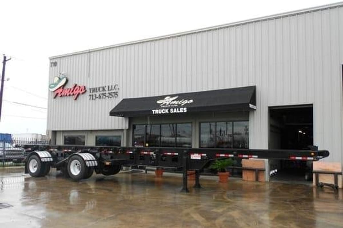Amigo Truck: Gray metal warehouse-style truck sales building with a black awning and signs, and a black trailer chassis parked out front