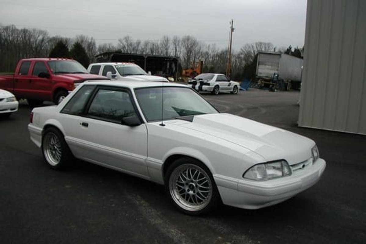 Delk Performance Parts: White coupe parked on an asphalt lot with pickup trucks, a trailer and a metal building in the background