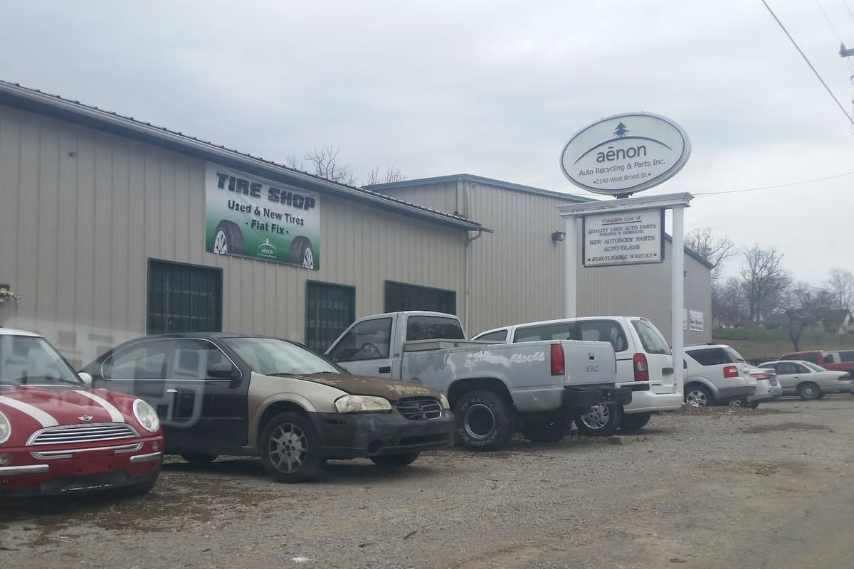 Aenon Auto Recycling and Parts: Tan metal shop building with a tire shop banner, a pole sign, and cars and trucks parked on a gravel lot