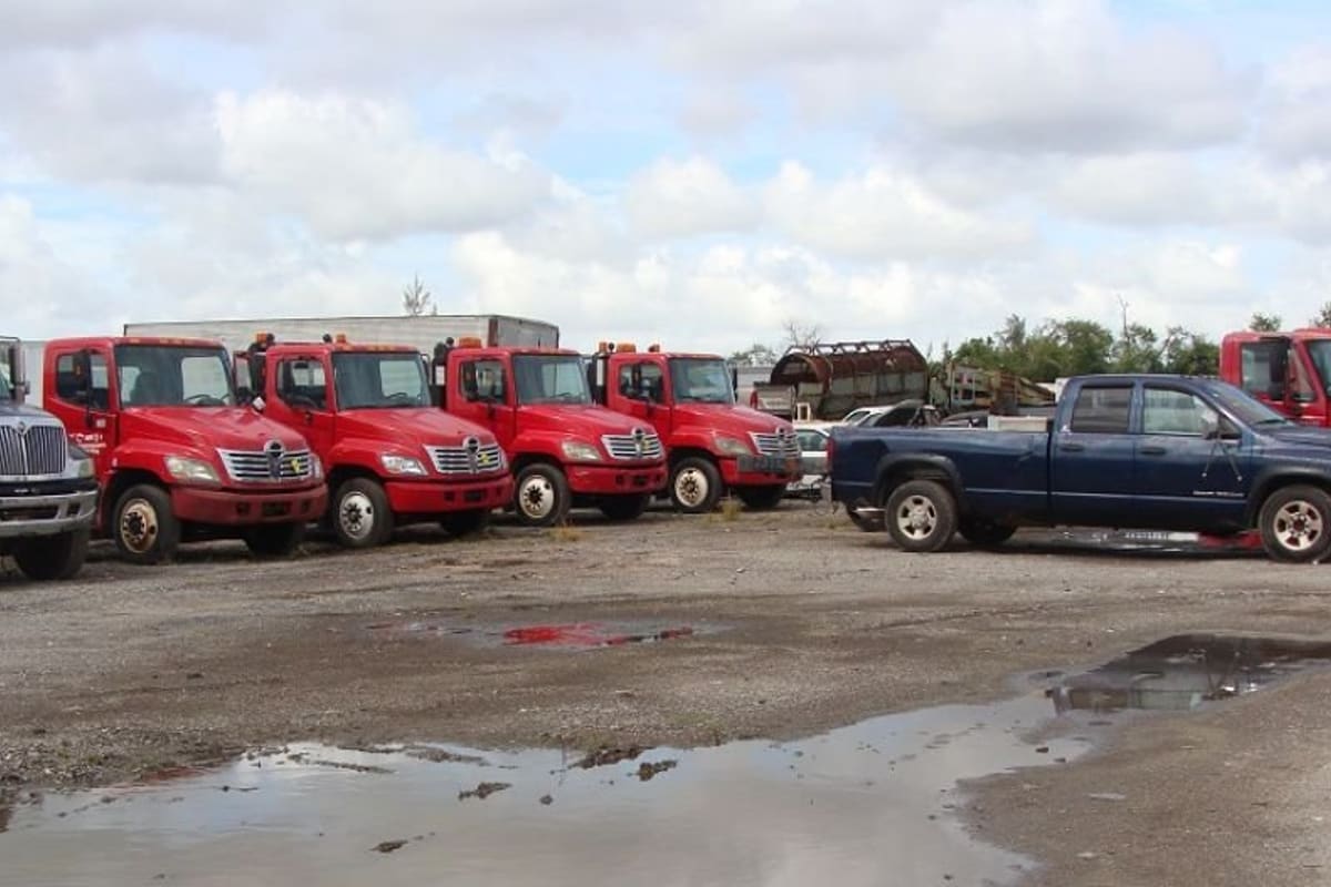 Big 3 Truck Salvage: Row of red and dark trucks and a blue pickup parked on a gravel salvage lot under a cloudy sky