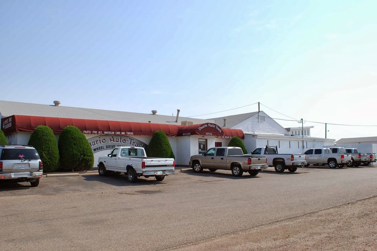 Daurio Auto Truck: White single-story office building with a red awning and trimmed shrubs, with pickup trucks parked in the lot under a clear sky