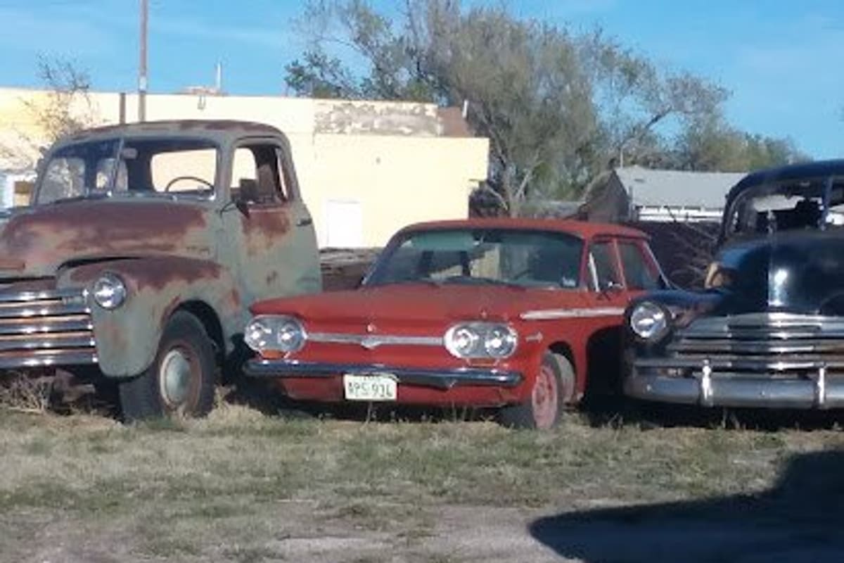 Amarillo Salvage: Rusty vintage pickup truck, red compact car and black classic car parked on grass in an outdoor salvage lot