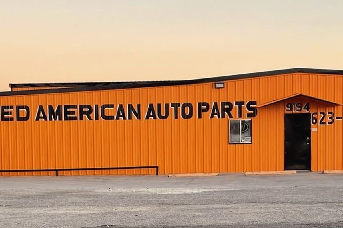 Allied American Auto Parts: Orange metal auto parts building with large black lettering and a front door, seen at sunset across a gravel lot
