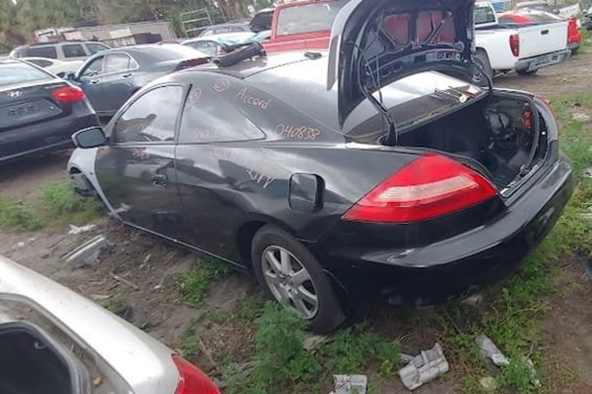 Bithlo Used Auto Parts: Salvage yard with a black coupe with open trunk, wrecked cars and parked vehicles on grassy dirt ground