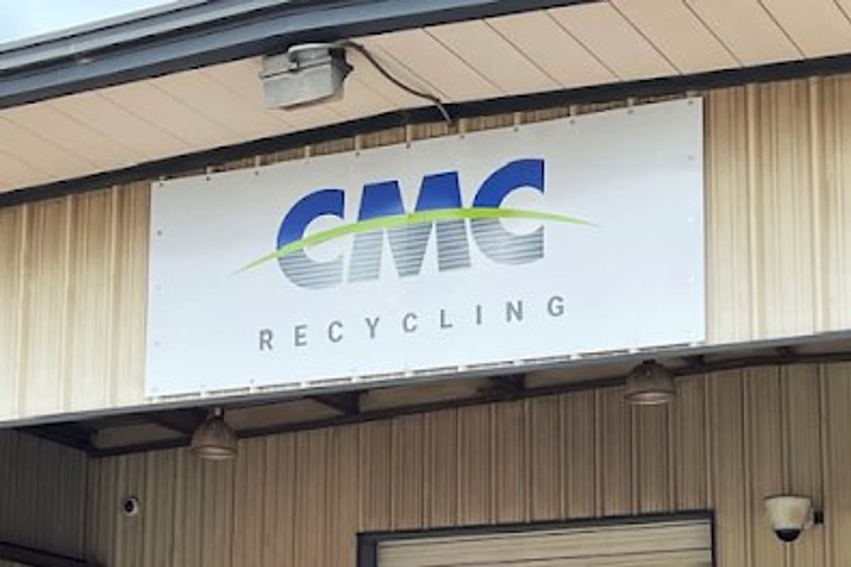 CMC Recycling: White recycling company sign mounted on a tan metal building wall under a roof overhang with lights and a camera