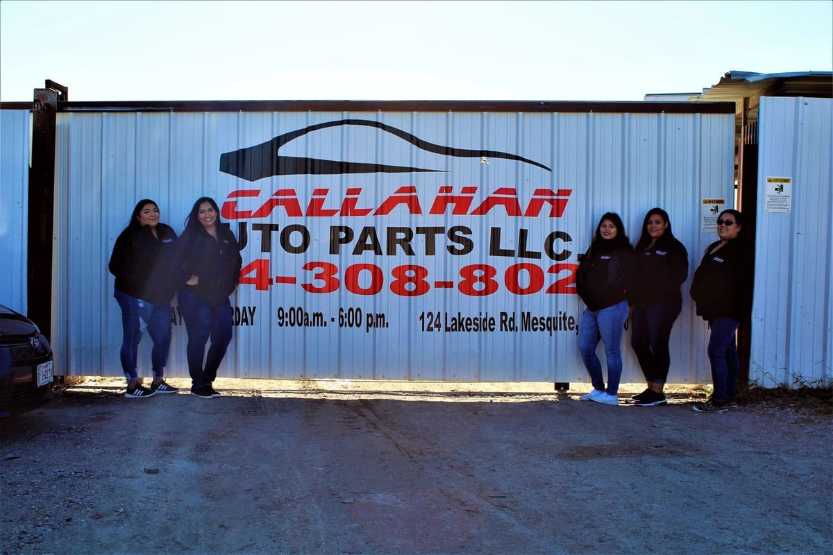 Callahan Auto Parts: Five women in dark jackets stand beside a corrugated metal gate with a painted auto parts sign, hours and address