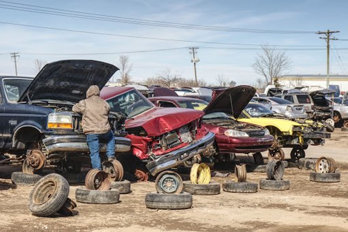 Hiller Auto Salvage: Salvage yard with wrecked cars, open hoods and loose tires on dirt ground, with a person working at a truck