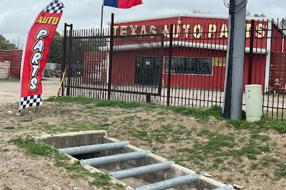 Texas Auto Parts: Red auto parts building with large yellow lettering behind a black iron fence, a Texas flag and an auto parts banner out front