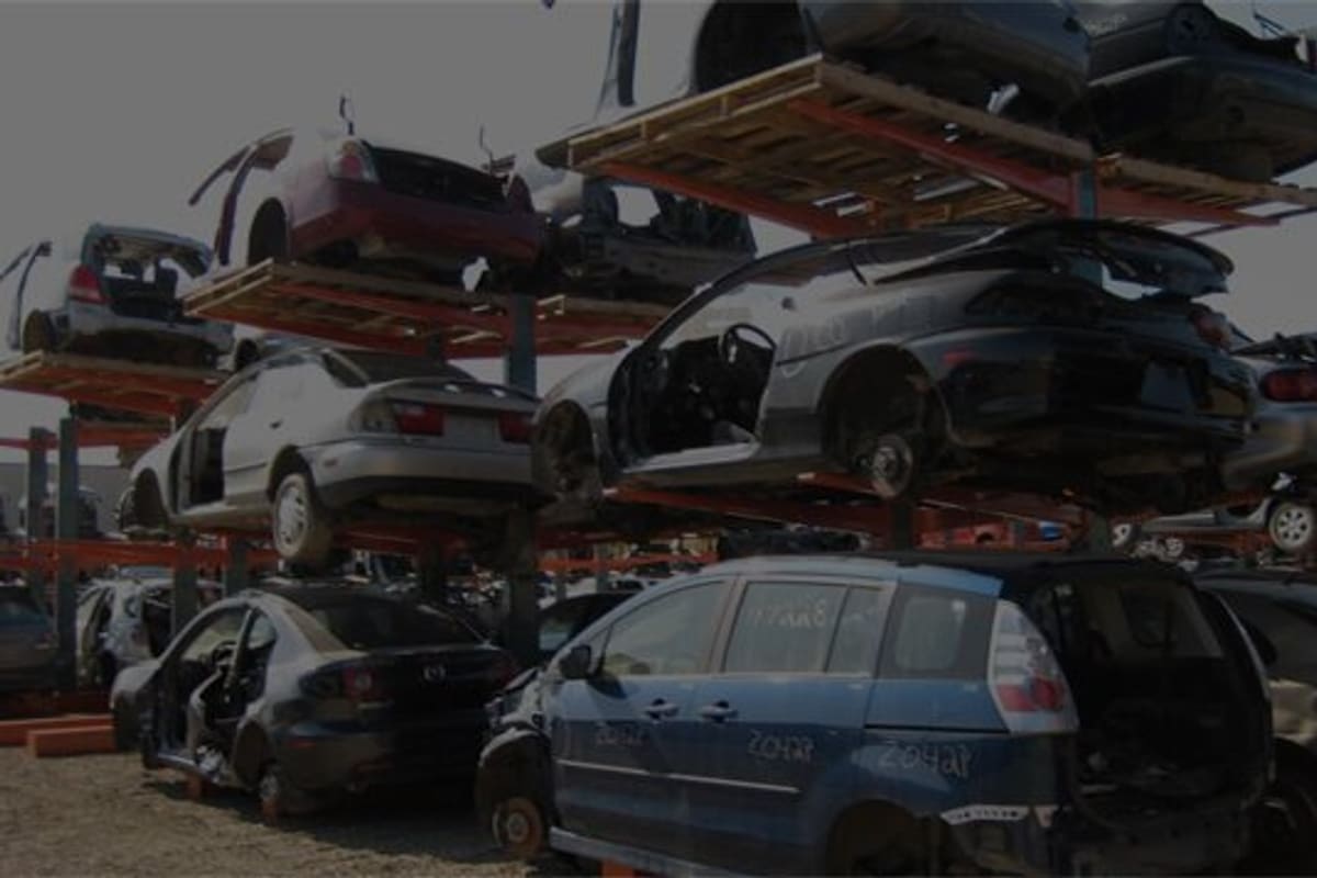 Nica Motors: Wrecked cars stacked on tall orange racks in a salvage yard, with more vehicles parked on gravel below