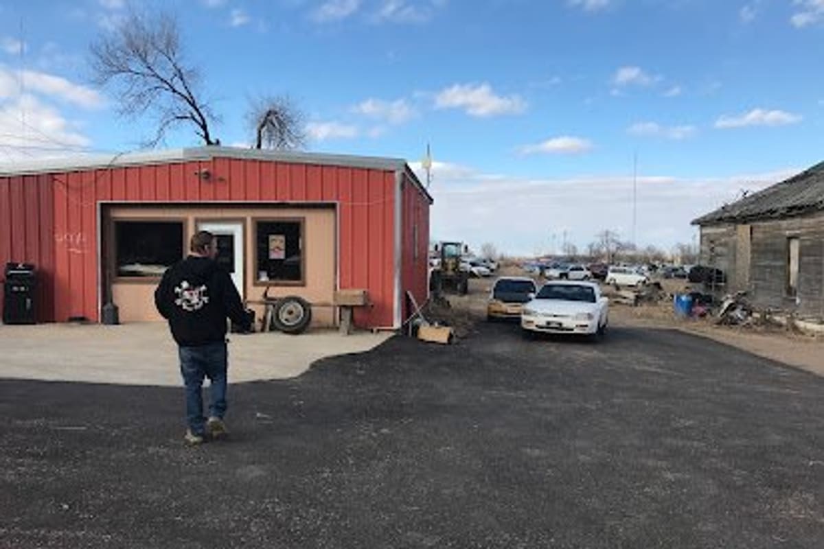 Clarks Auto Salvage: Red metal office building beside a gravel lot with cars parked along a dirt road and an old wooden shed nearby
