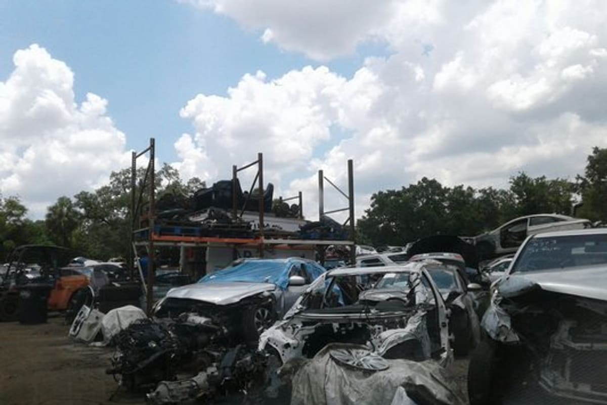 TMT Salvage: Wrecked and dismantled cars in a salvage yard with a tall storage rack and trees under a cloudy sky