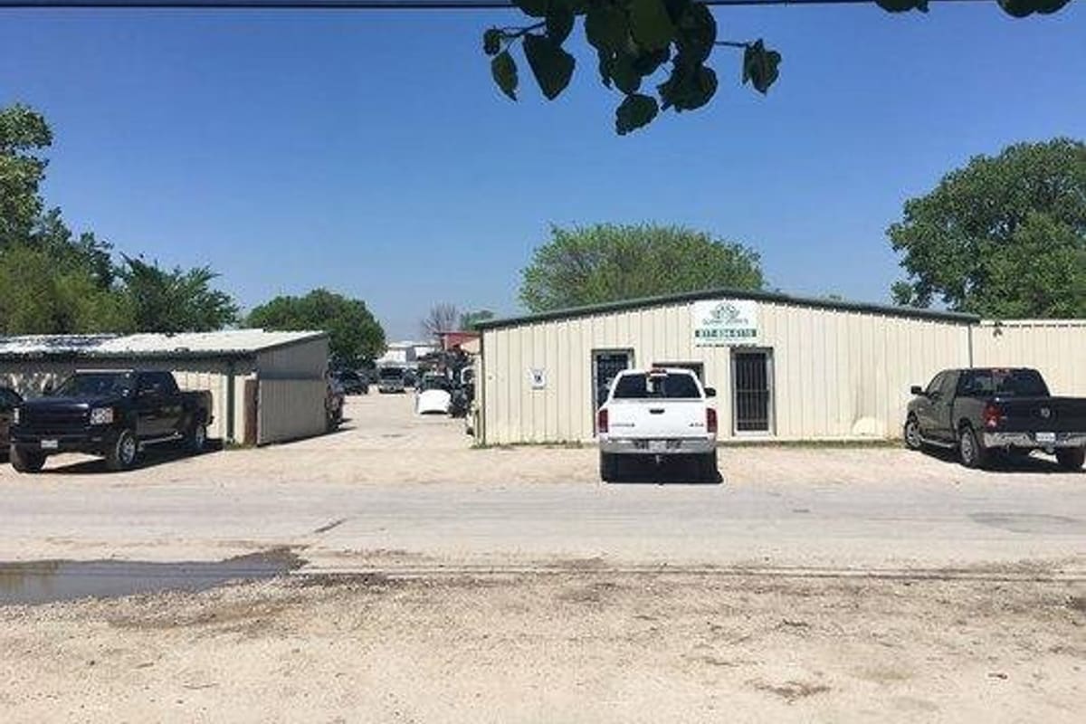 AAA Packs: Metal office building with a small sign and pickup trucks parked on a gravel lot beside a covered shed on a sunny day