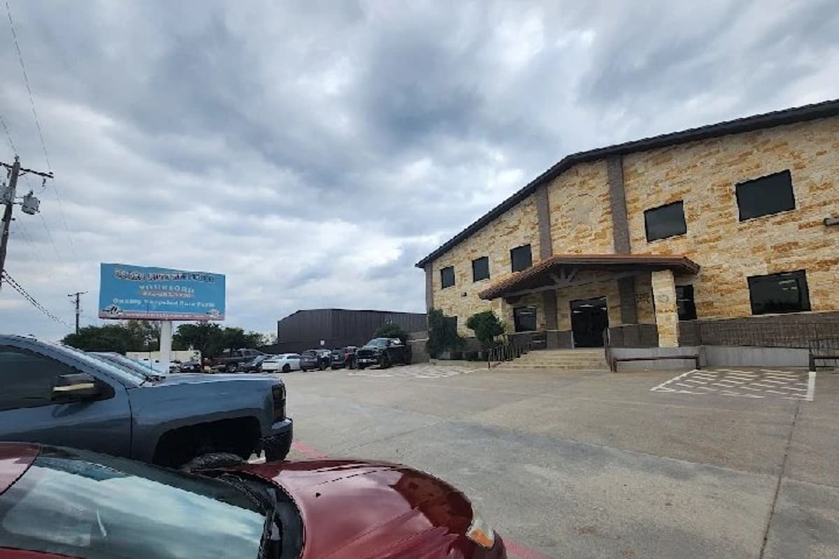 Yourford/Texas Auto Recyclers: Two-story stone office building beside a paved parking lot with cars and a roadside sign under a cloudy sky
