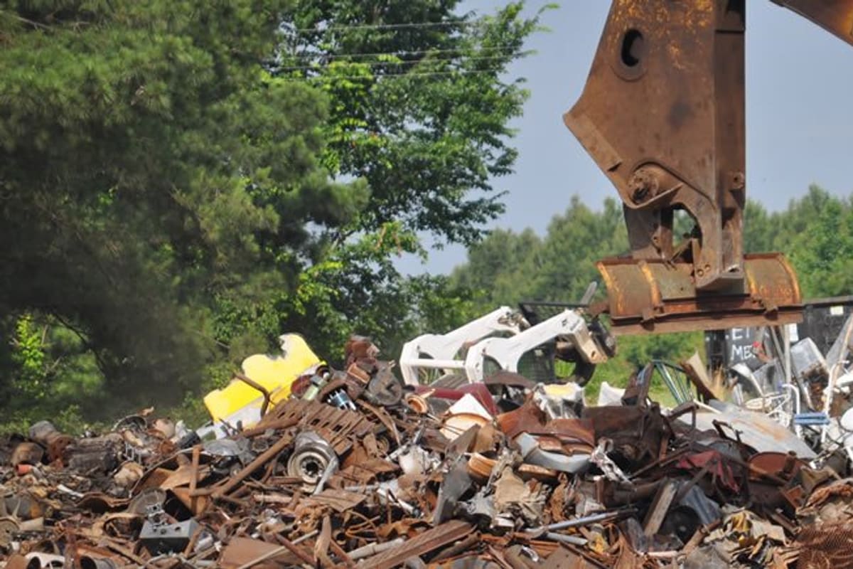 East Texas Recycling: Yellow excavator with a grapple loading a large pile of scrap metal at a salvage yard surrounded by trees