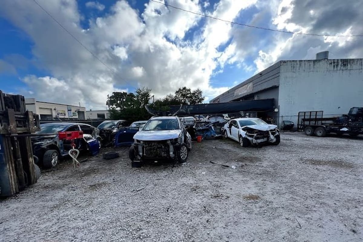 Warner Salvage Auto Parts: Gravel salvage yard with wrecked cars parked in front of a warehouse building under a cloudy sky
