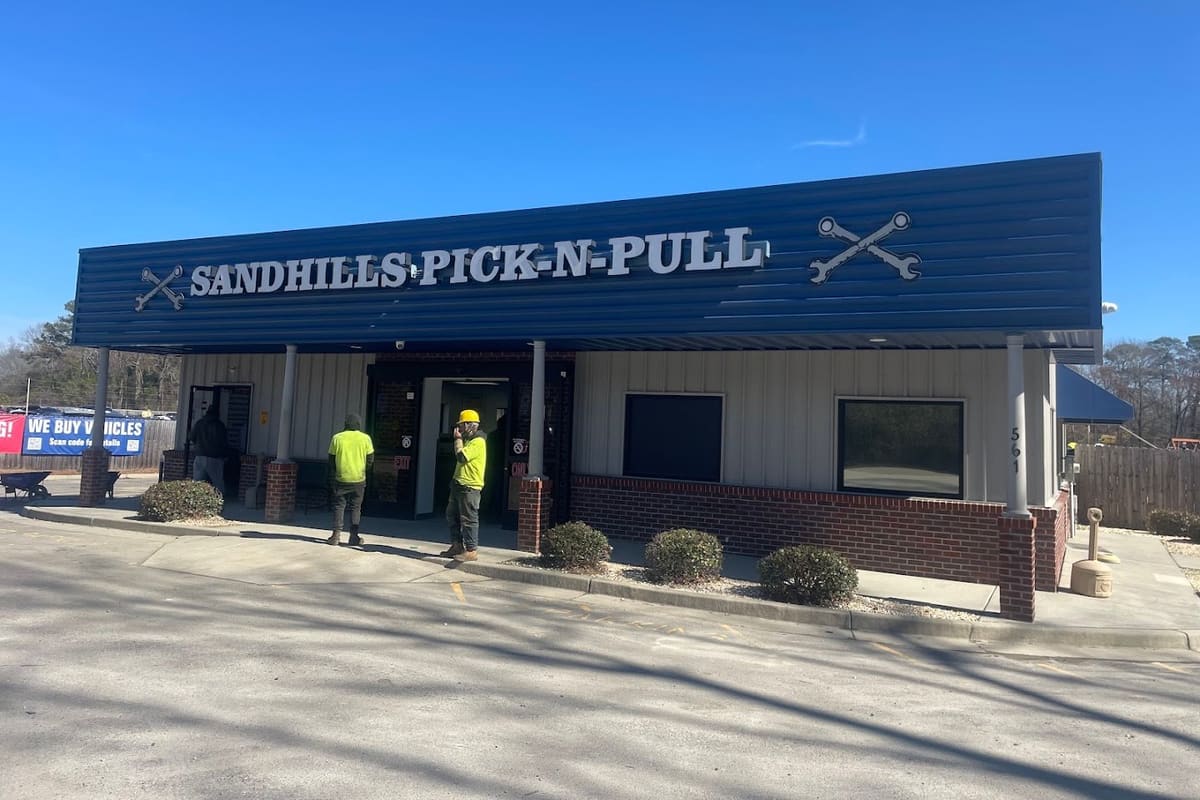 Sandhills Pick N Pull: Single-story self-service junkyard office with a blue metal roof, brick base, crossed wrench signs and shrubs, under a clear blue sky