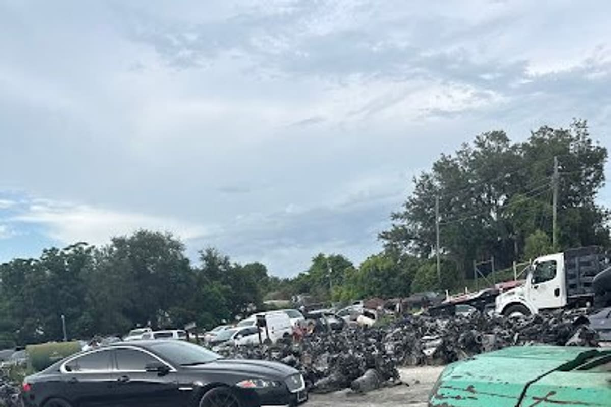 Central Florida Auto Recycling: Gravel salvage yard with a black sedan, a green forklift, and piles of scrap vehicle parts under a cloudy sky