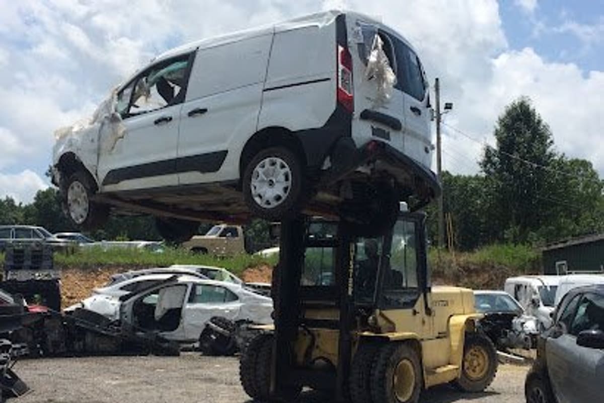Smithville Auto Salvage: Forklift lifting a white van high above a gravel salvage yard with wrecked cars parked around under a cloudy sky