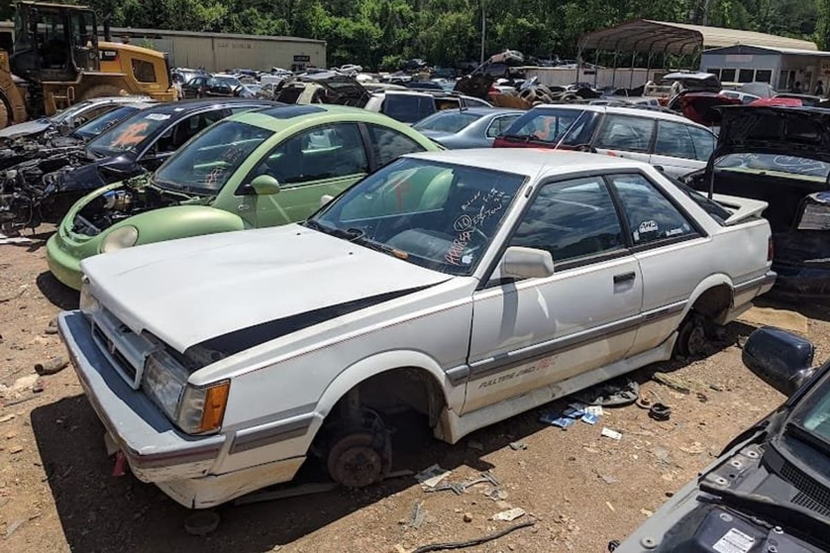 Lambert's Pick-a-Part: Rows of wrecked cars parked on dirt in a salvage yard, with a white coupe and a green car in front and a loader behind