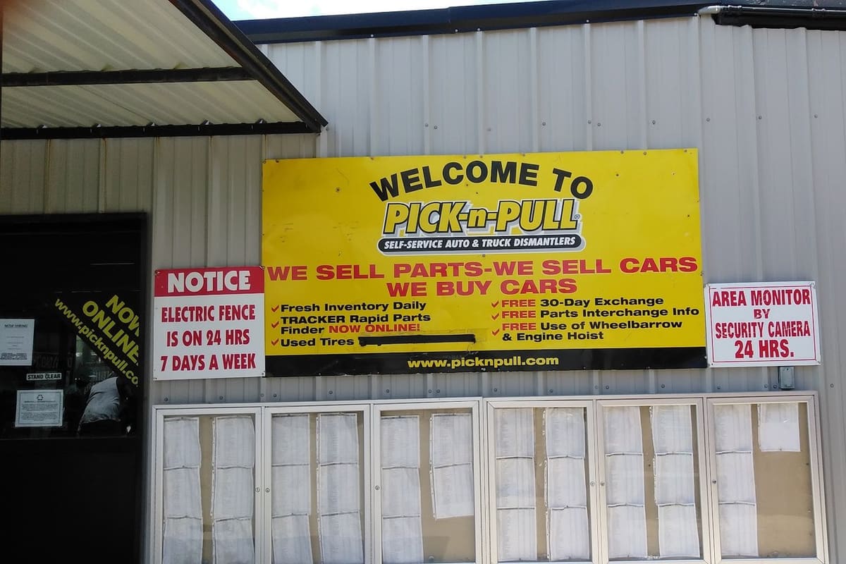 Pick-n-Pull - Dallas South Central: Yellow welcome sign on a metal building listing parts and car buying, with notices about an electric fence and security cameras