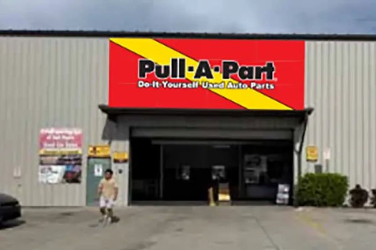Pull-A-Part Fort Myers: Large metal warehouse-style building with a red and yellow sign above an open garage-style entrance and a parking area in front