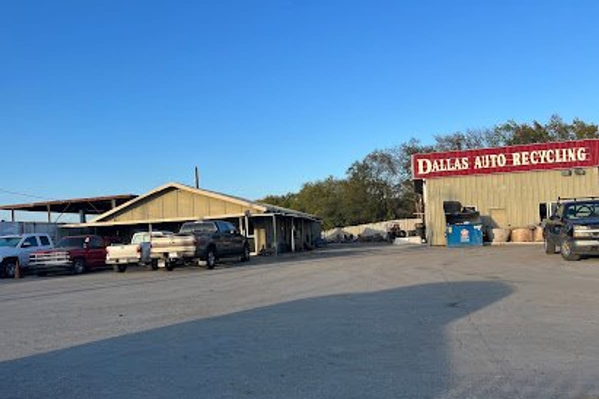 Dallas Pick a Part: Wide daytime view of a gravel lot with pickup trucks parked beside a tan building and a red sign reading Dallas Auto Recycling