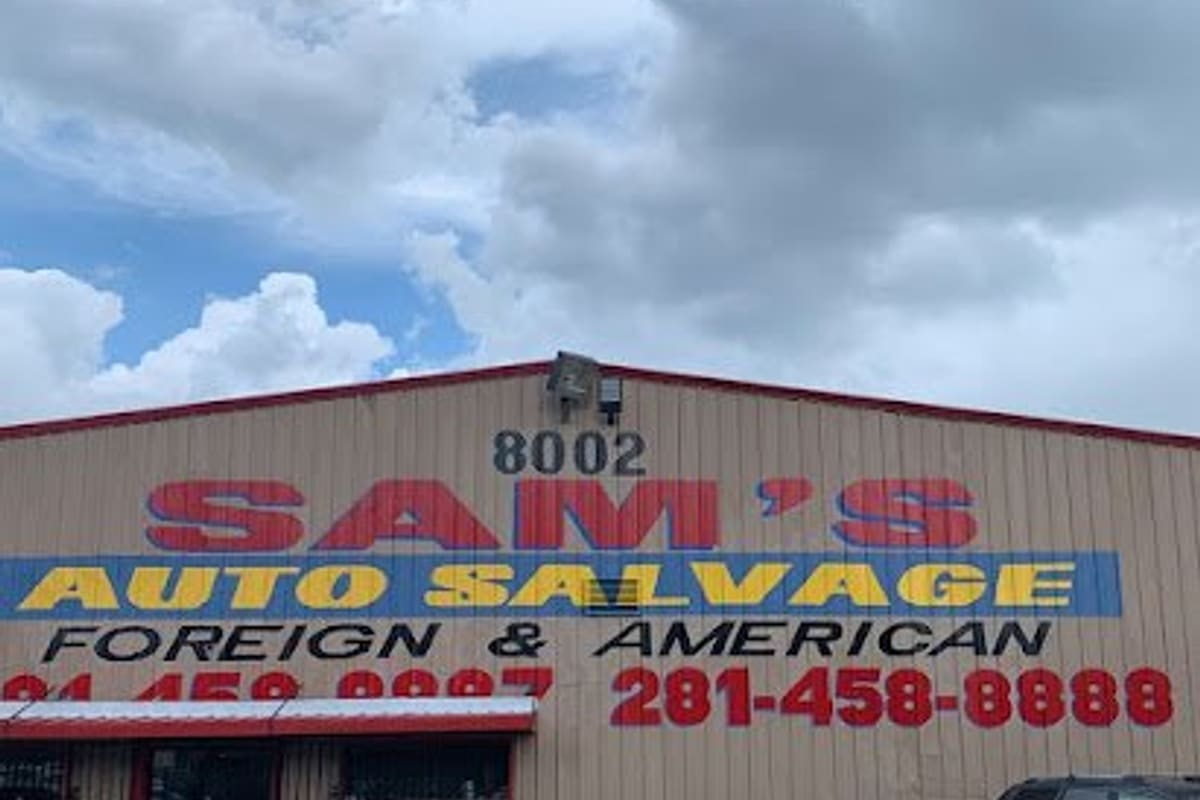 Sam's Auto Salvage: Tan metal warehouse building with a large painted auto salvage sign and phone number, with cars parked in the gravel lot in front