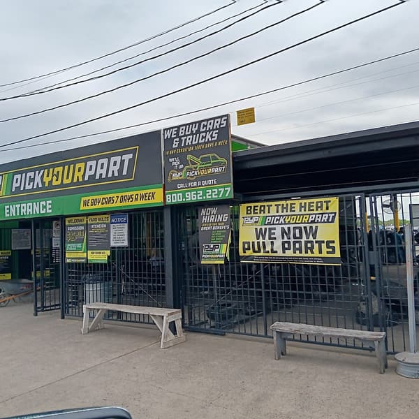 Pick Your Part - Houston Wallisville: Gated entrance and exit of a self-service auto salvage yard with green and black signs, benches and wheelbarrows out front